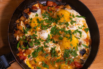 Close-up of fried chicken eggs, tomatoes, eggplant with seasoning and fresh parsley in a frying pan. Cooking process, selective focus. Homemade food for breakfast
