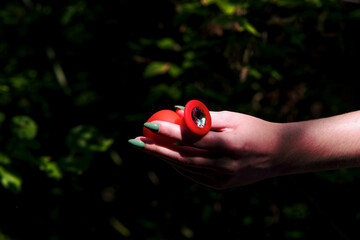Woman's hand holds a red anal plug against a background of green bushes in the forest. Sex toys,...