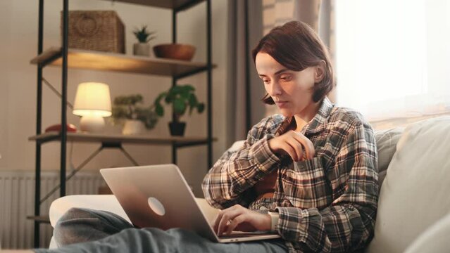 Portrait of pretty young woman freelancer having distance remote work typing browsing scrolling products in internet online store on laptop computer and eating cookie from pocket at light home indoors