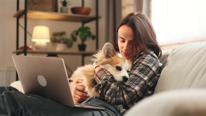 Pretty young woman take a break from laptop work relaxing with fluffy corgi dog hugging sitting on couch at cozy home Smiling girl having leisure time with her little pet at home Best friends