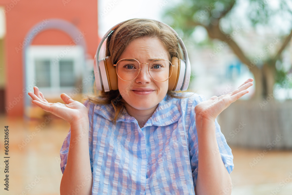 Poster Young redhead girl headphones at outdoors having doubts while raising hands