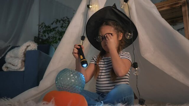 Cute happy child girl in top hat sits in tent, plays with magic lantern with glass ball.child fantasizes, dreams of witchcraft, fulfillment of desires.Happy child at home playing in room for christmas