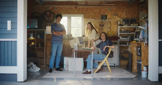 Diverse Team Of Three Young Startup Company Founders In Retro Garage With Old Desktop Computer. Male And Female Entrepreneurs Looking At Camera And Smiling, Developing Online Service In Nineties.