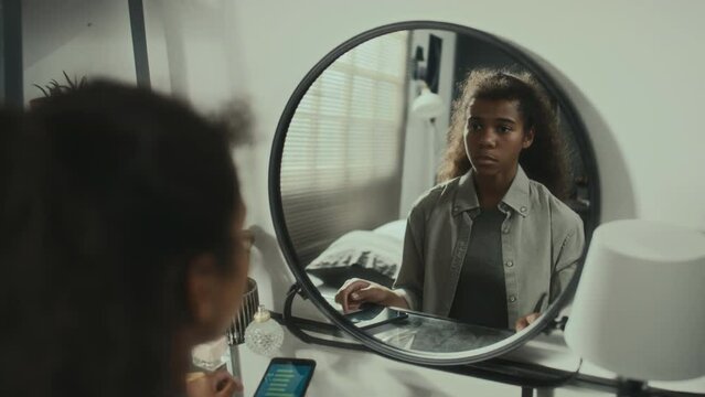 African American teen girl sitting at home, reading humiliating message on phone, looking at her reflection in mirror and getting upset about cyberbullying