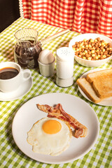 Fried egg with roasted bacon, black coffee, toasts on checkered tablecloth. Retro American breakfast. Hard shadow.
