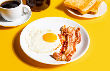 Fried egg with roasted bacon, black coffee, toasts on yellow background. Retro American breakfast. Hard shadow.
