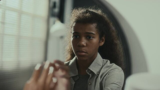 Black teen girl sitting at home, reading embarrassing message on mobile phone, looking in mirror and hating her reflection