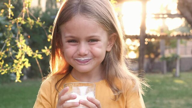 Happy emotional smiling adorable child girl hold glass of milk and drink on nature outside sunny summer morning. Kid open mouth with mustache of milk on lips. Calcium for good children health