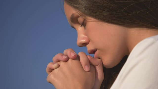girl closed her eyes, praying to heaven.Hands folded in prayer faith concept.concept of peace, hope, dreams. Spiritual face of young white girl praying with hope, eyes closed, hands folded in prayer.