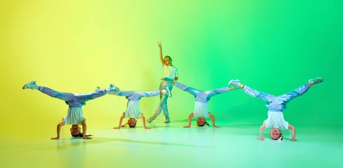Fotobehang Dansschool Happy children dancing on head. Group of little cute girls dressed in fashion, stylish outfit dancing in choreography class isolated on green-yellow gradient background in neon light.