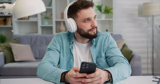 Young calm man 25s wear shirt headphones, sit for table hold and use mobile cell phone look aside listen music stay at home rest relax spend free spare time in living room, indoors