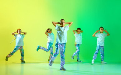 Group of little cute girls wearing style clothes with trendy, creative hairstyle hold hands in choreography class against studio background in yellow-green neon light. - obrazy, fototapety, plakaty