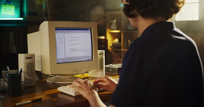 Back View Of Caucasian Male Software Engineer Programming on Old Desktop Computer In Retro Garage. Focused Man Starting Tech Startup Company In Nineties. Coding Innovative Online Service At Home.
