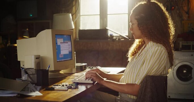Side View Of Hispanic Female Engineering Student Writing Academic Thesis On Old Desktop Computer In Retro Garage. Future Engineer Focused On Innovative Project For University Of Technology In Nineties