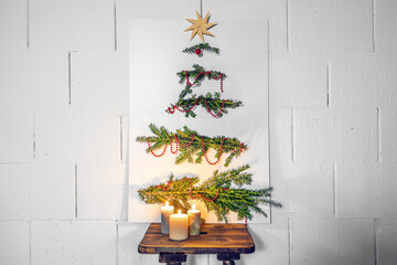 Fototapeta na wymiar Homemade Christmas tree alternative, white cardboard plate with fir branches and decoration leaning against the wall on an old wooden stool with candles, copy space