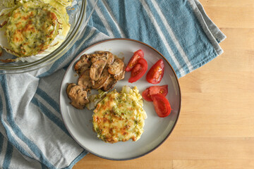Gratinated Chinese cabbage casserole with mushrooms and tomatoes, healthy low carb diet meal on a blue kitchen towel and a wooden table, copy space, top view from above