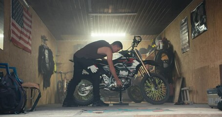 A male mechanic with a short haircut in a gray T-shirt removes his bike from the jack and takes it out of his garage workshop. Male mechanic completing motorcycle repair - Powered by Adobe