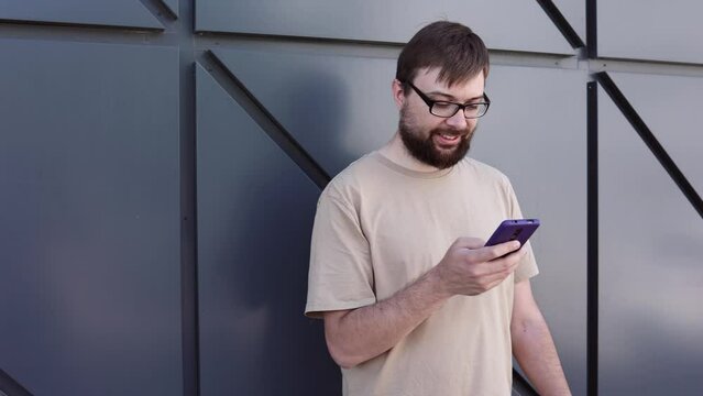 Handsome man with a beard and glasses uses a phone against the background of a wall. Drag. Use of technology, online messaging, smartphone, blogger