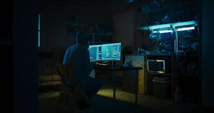 Home Garage Late At Night: Evil Male Hacker Wearing Hoodie Breaks into Data Servers, DDOS Attack, Phishing Attack, Malware. Man Writing Code For Trojan Horse Virus. Darknet Cyber Crime Concept.