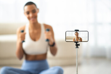 Smiling young latin lady with dumbbells in sportswear shooting video fitness blog on smartphone