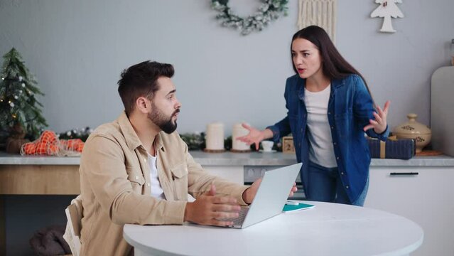 Upset angry wife addresses husband engrossed in laptop during Christmas in festive kitchen, portraying financial strains causing familial disputes. Intimate household quarrel during New Year holiday