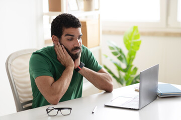Sad tired young middle eastern guy blogger sit at table, look at laptop, has work problems