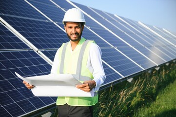 Portrait of Young indian man technician wearing white hard hat standing near solar panels against blue sky.Industrial worker solar system installation, renewable green energy generation concept.