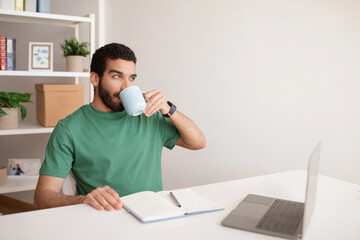 Positive millennial arab guy planning startup business at computer, drink cup of tea, enjoy coffee break