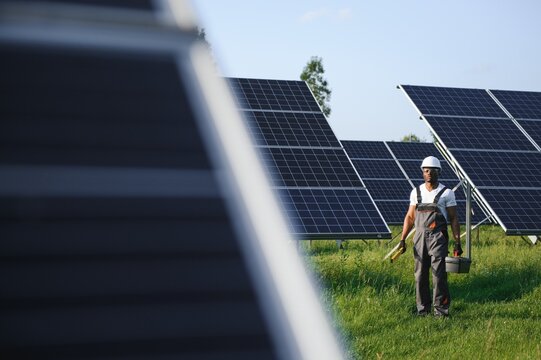 Competent energy engineer in grey overalls and white helmet checking solar panels while walking on field. African american man carrying clipboard and container with instruments