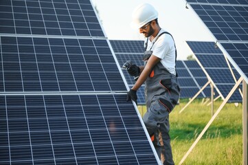 Male arabian engineer in helmet and brown overalls checking resistance in solar panels outdoors. Indian man working on station.
