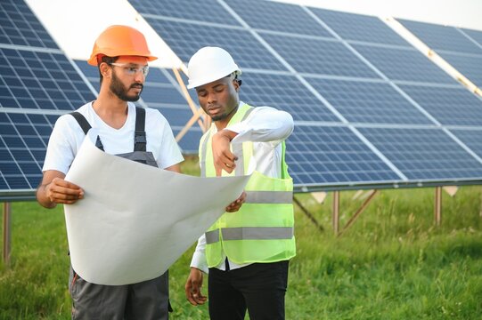 Group of multi ethnic people in safety helmets standing at solar station. Two engineers and technician examining plan of panels outdoors.