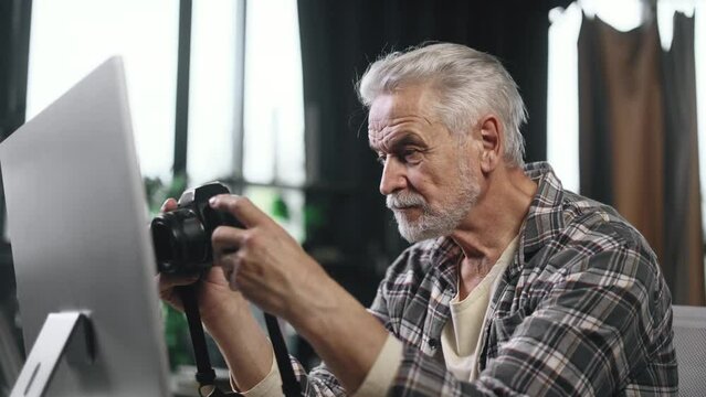 Portrait of handsome gray haired senior man photographer hold digital camera looking at screen choosing photos for editing while sitting in front of computer at home workplace