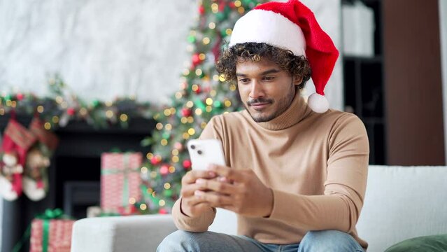 Young man in santa hat uses smartphone while sitting on sofa at home during winter New Year Christmas holidays. A handsome male is writing a greeting message, chatting with friends or shopping online