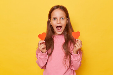 Shocked amazed brown haired little girl wearing pink sweatshirt isolated over yellow background receives red hearts on Valentines Day from classmate being astonished and surprised.