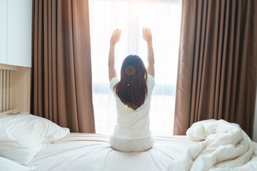 happy woman stretching on bed after wake up, young adult female rising arms and looking to window...