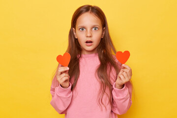 Shocked scared brown haired little girl wearing pink sweatshirt isolated over yellow background holding two big red hearts looking at camera with eyes and open mouth.