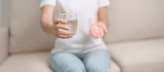 woman hand holding medicine painkiller pill and water glass on the sofa at home, taking for...