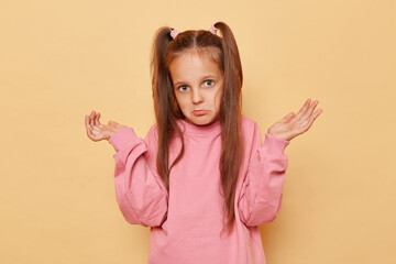 Puzzled uncertain little girl with ponytails wearing pink sweatshirt posing isolated over beige background shrugging shoulders looking with pout lips doesn't know answer.