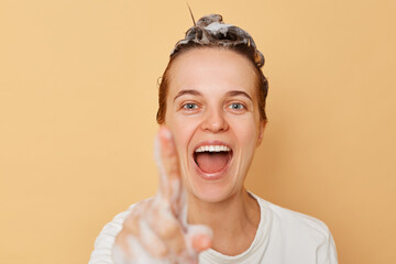 Beautiful cheerful joyful woman washes hair applies shampoo taking shower isolated over beige background showing foam on her finger click invisible button.