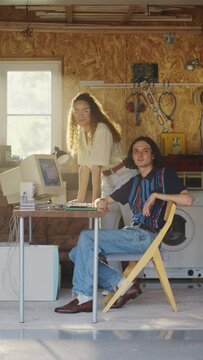 Vertical Screen: Caucasian Male Software Developer And Hispanic Female Designer Looking At Camera And Smiling In Retro Garage With Old Computer. Startup Founders Working On Online Service In Nineties.