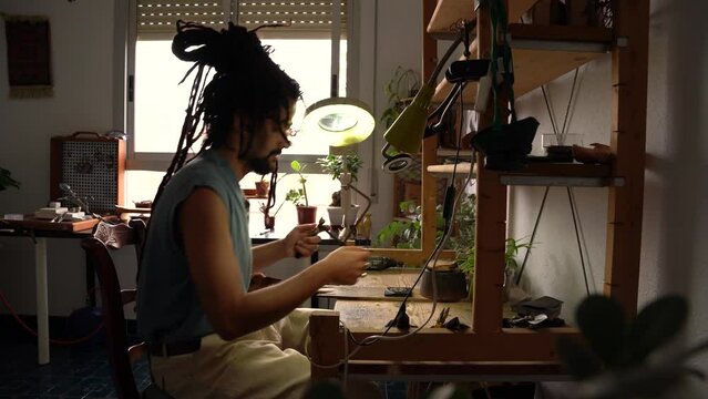 Young Rastafarian jeweler in a goldsmith's workshop, sitting at his jeweler's bench cutting out pieces with a hacksaw.