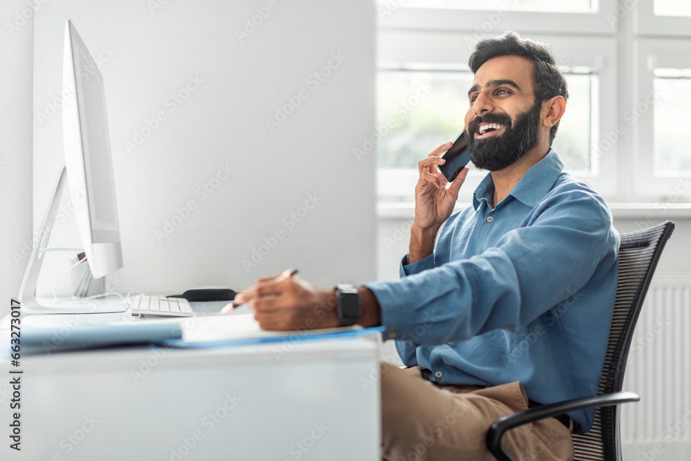 Poster positive thoughtful indian businessman talking on cellphone and taking notes while working on comput