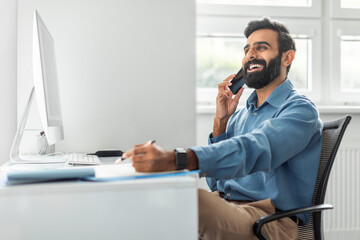 Positive thoughtful indian businessman talking on cellphone and taking notes while working on computer in office