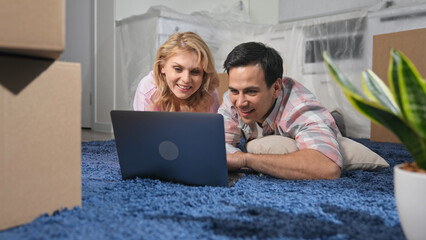 Happy family lying carpet use laptop. Couple choose new furniture. Spouses moving house. Real...