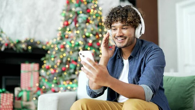 Young Handsome Man In Wireless Headphones Listening To Music While Sitting On Sofa At Home During Winter New Year Christmas Holidays. Happy Guy Turns On A Playlist On His Smartphone And Enjoys Dancing