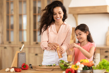 Mother and daughter kid adding spices to salad in kitchen