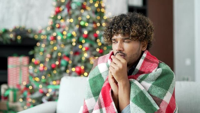 A young handsome man covered with a blanket froze sitting on sofa in living room at home during winter New Year Christmas holidays. Male cold indoors. Guy feels discomfort due to low temperature