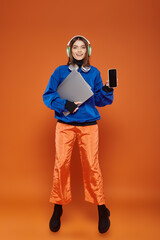 happy young woman in headphones and autumnal outfit holding smartphone and laptop, cyber monday