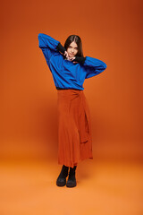 autumn fashion, attractive woman in skirt, blue sweatshirt and boots standing on orange backdrop