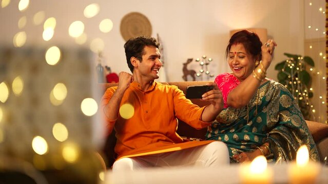 Indian son and mother watching cricket match on mobile and clap with both hands,while both are sitting on the sofa.well decorated home,A hindu festival